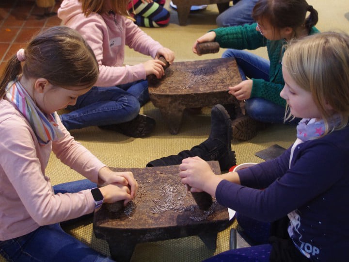 Kinder reiben Kakaonibs auf mexikanischen Reibsteinen zu Kakaomasse